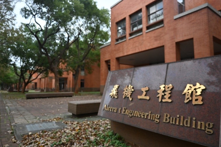 實習工廠戶外場景，前景是一個科系標示牌，上面寫著「飛機工程館」及英文名稱，背景為兩層樓的紅磚建築，是一個培養直升機維修人才的學術機構。
